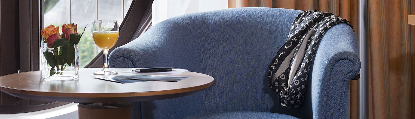 Detail of the hotel room  Cozy seating area in the Classic Room of Maritim Hotel Fulda with blue armchair, round window, flowers, and orange juice.