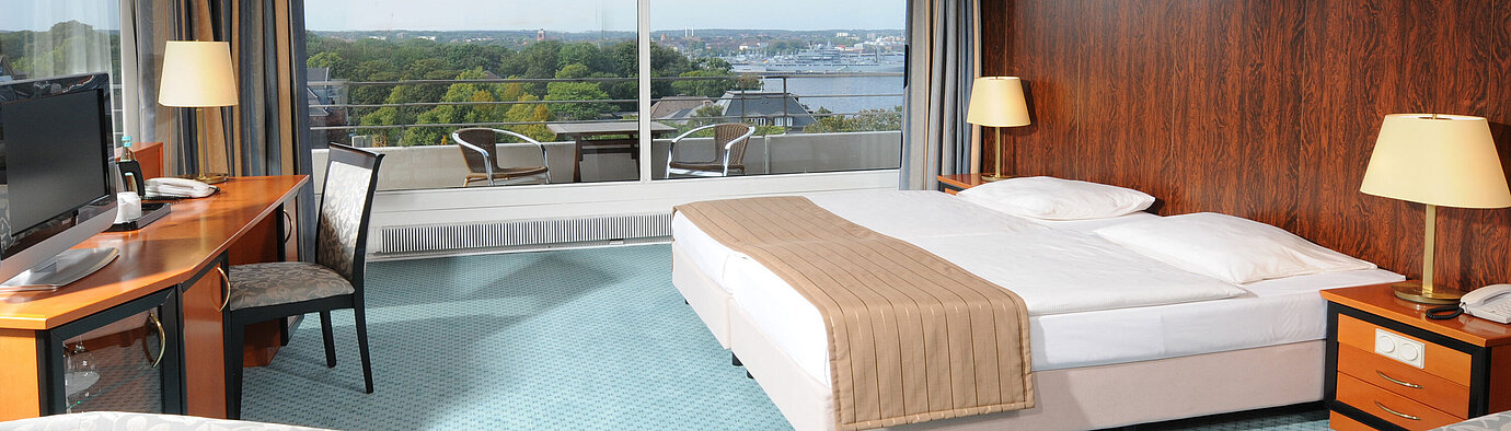 Spacious hotel room with a double bed, seating area and wide views of Kiel through the large window front.