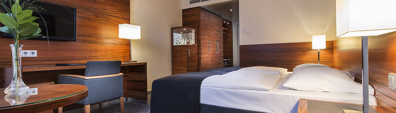Furnished hotel room at the Maritim Hotel Munich with a comfortable bed, workspace, TV, and wood paneling.