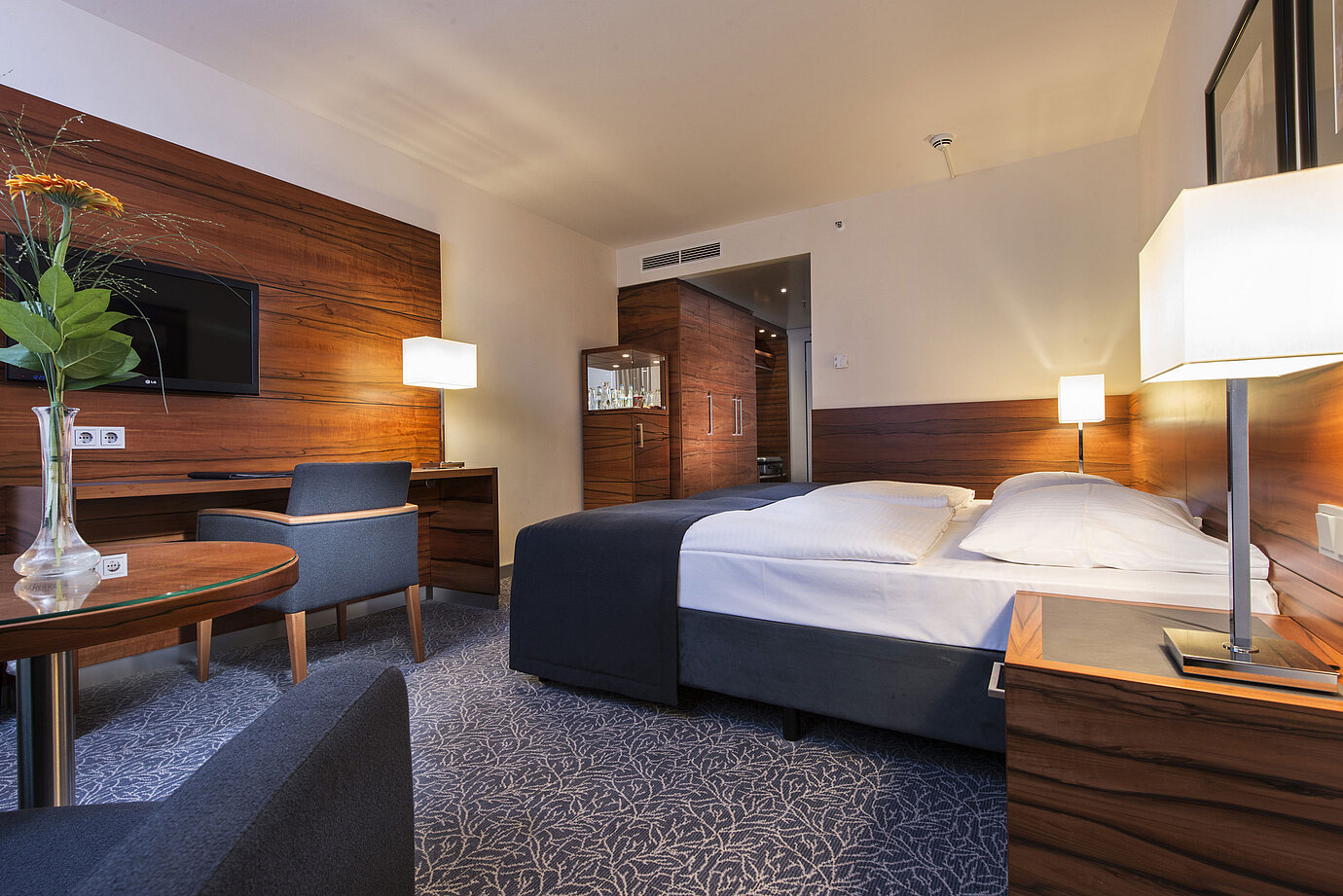 Furnished hotel room at the Maritim Hotel Munich with a comfortable bed, workspace, TV, and wood paneling.