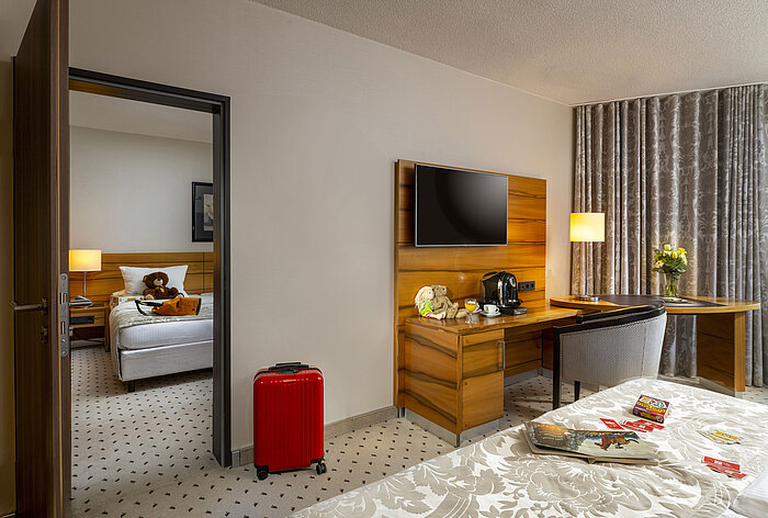 Family room at Maritim Hotel Bad Homburg with separate kids' room, teddy bears, and red suitcase.