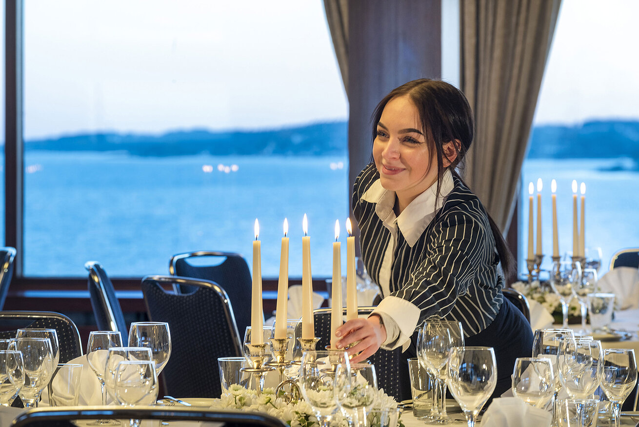 Hall Maritim in the Maritim Hotel Kiel, a member of staff lights candles on a festively laid table, sea view in the background.