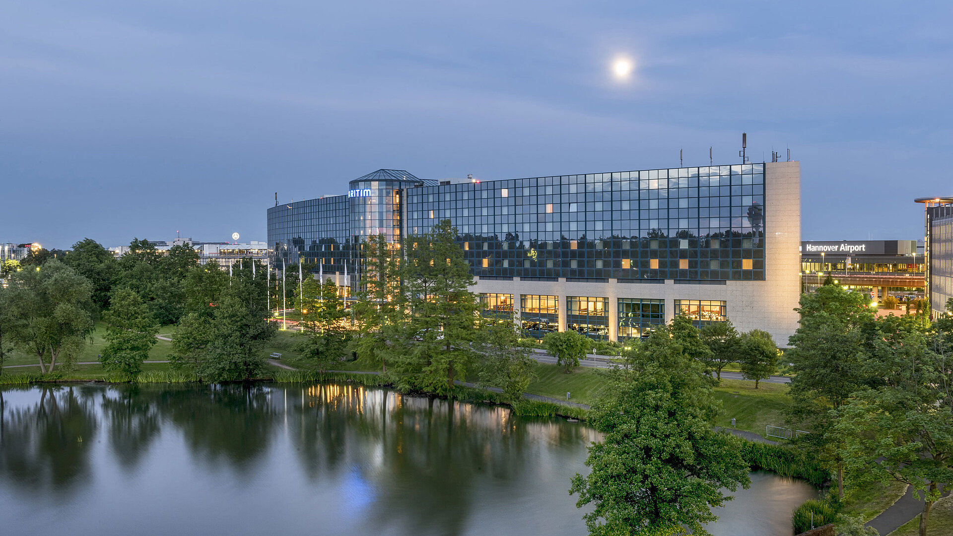 Illuminated Maritim Hotel Hannover with a glass facade and lake reflection at dusk.