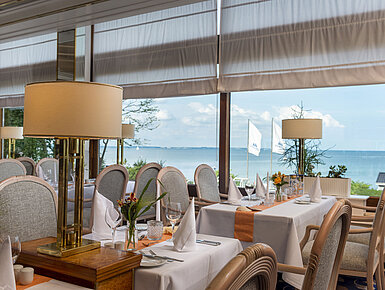 Elegant restaurant with set tables and a sea view, decorated with lamps and flowers for a fine dining experience.