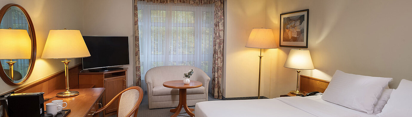 Classic room with double bed, desk, TV, seating area and large window in a bright interior design.