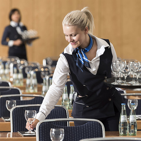 A service staff member at the Maritim Berlin sets up a conference room, carrying a tray of glasses and placing water bottles.