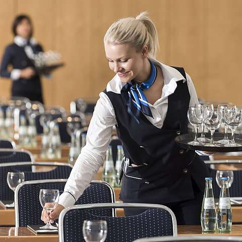 A service staff member at the Maritim Berlin sets up a conference room, carrying a tray of glasses and placing water bottles.