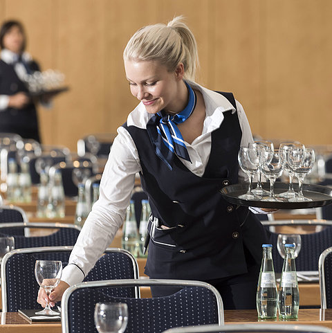 A service staff member at the Maritim Berlin sets up a conference room, carrying a tray of glasses and placing water bottles.