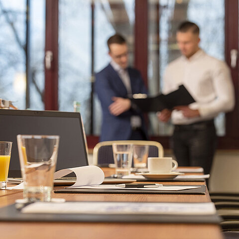 Conference room with a laptop, drinks, and documents, two businessmen in the background discussing papers.