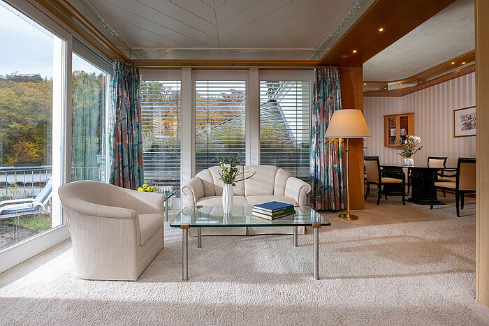 Bright hotel suite living area with glass table, cosy armchairs and large windows offering a view.