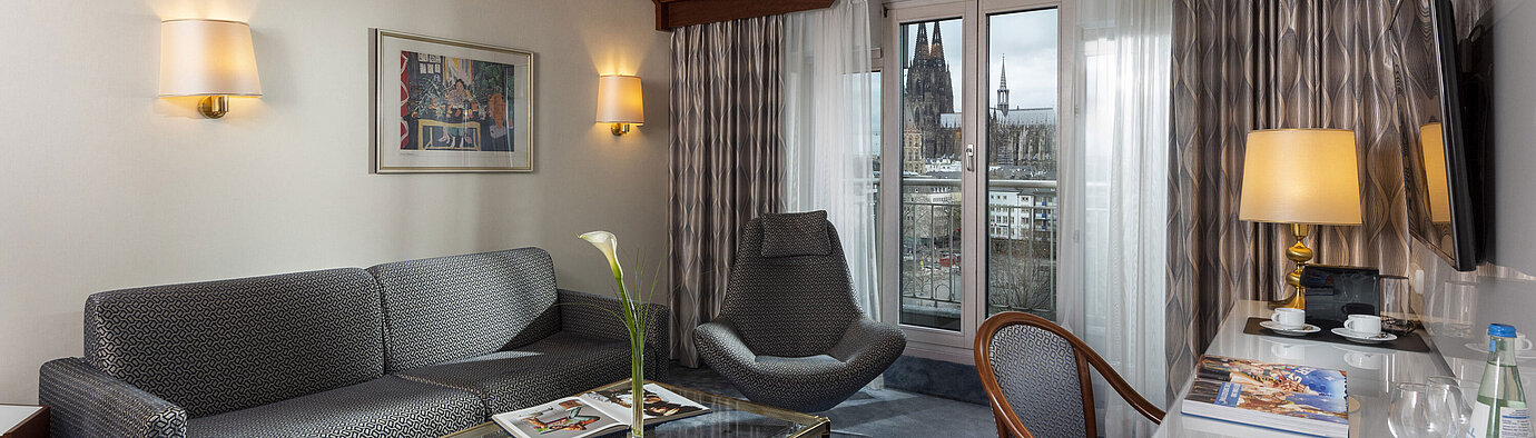 Junior Suite Junior Suite at Maritim Hotel Cologne with a view of Cologne Cathedral, stylish sofa, chair, and desk with reading material.
